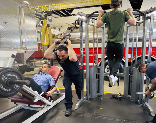 three firefighters working out in a gym located inside a fire station. One firefighter is performing leg presses, another is doing cable tricep extensions, and the third is doing pull-ups. A fire truck is visible in the background